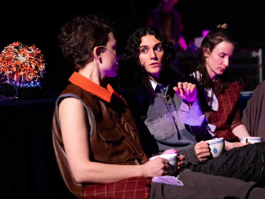 Drei Personen sitzen nebeneinander und halten Teetassen. Die mittlere Person blickt zur Seite, während die andere spricht. Das Licht mischt warme und violette Farbtöne; im Hintergrund sind technische Geräte sichtbar.