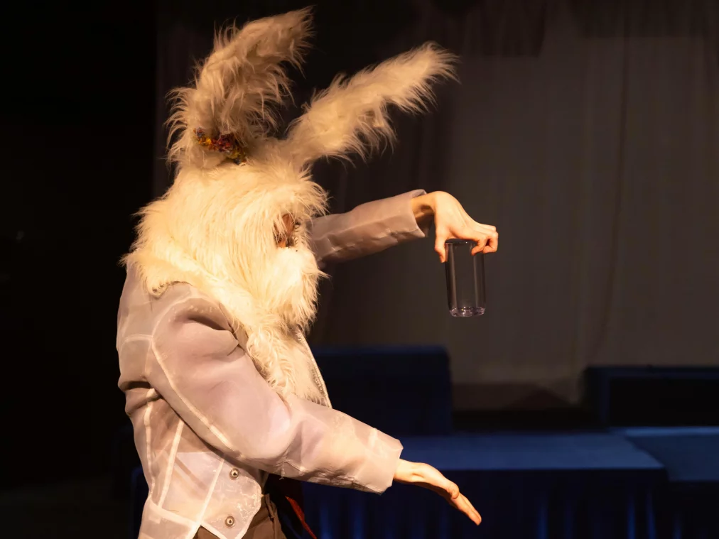 Eine Person trägt eine große weiße Tiermaske mit langen Ohren und hält einen dunklen Becher in der Hand. Sie steht seitlich im warmen Bühnenlicht vor einem hellen Vorhang.©MayraWallraff-160