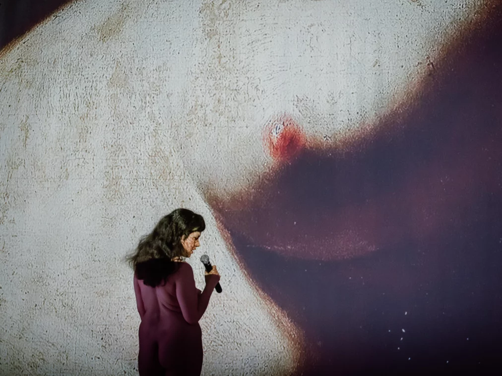 One person stands on a stage holding a microphone. Behind the person, a large-scale projection of a body fills the background. The person looks downward while the projected image stretches across the wall at a much larger scale.