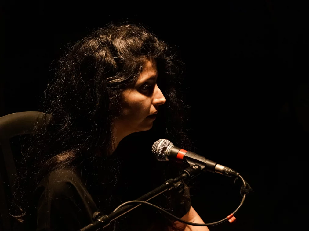 Eine Person sitzt im dunklen Bühnenraum vor einem Mikrofon. Das Gesicht der Person wird von einem warmen Spotlicht beleuchtet, während der Hintergrund schwarz bleibt.