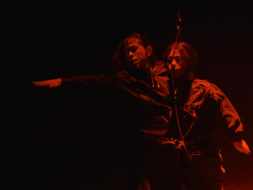 Zwei Personen stehen eng nebeneinander in dunklem Raum und werden von rotem Licht beleuchtet. Eine Person streckt einen Arm seitlich aus, während beide Körper nah beieinander stehen.