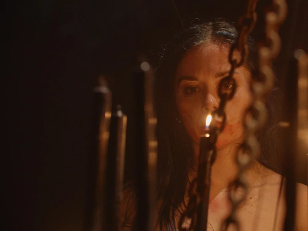 Eine Person steht zwischen mehreren brennenden Kerzen, die auf Metallhalterungen befestigt sind. Ketten hängen im Vordergrund der Szene. Warmes Kerzenlicht beleuchtet das Gesicht der Person.
