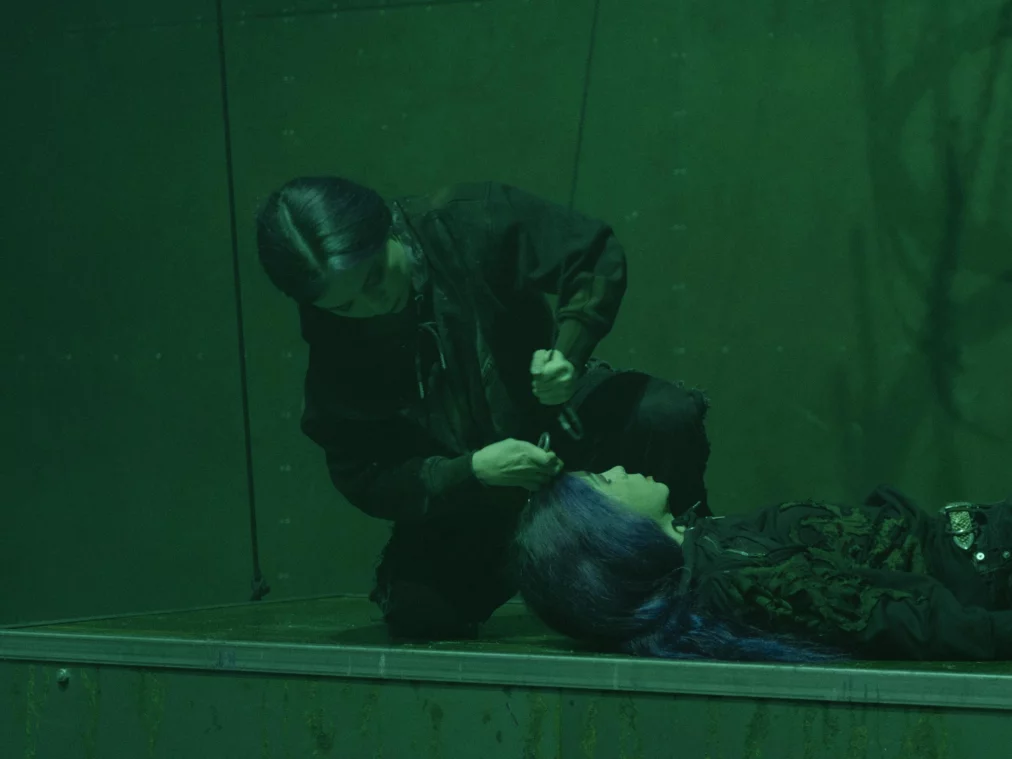 Eine Person kniet neben einer zweiten Person, die auf einer metallischen Plattform liegt. Die kniende Person hält ein Werkzeug in der Hand und arbeitet am Haar der liegenden Person. Die Szene ist in grünes Licht getaucht.