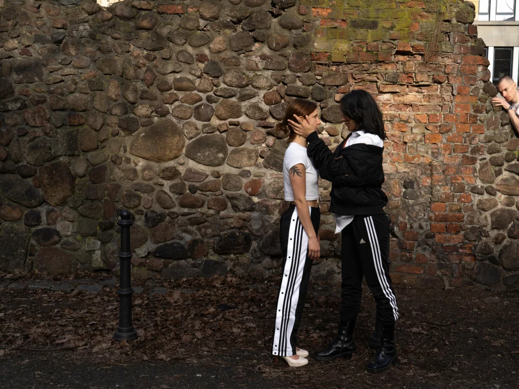 Drei Personen stehen im Freien vor einer alten Steinmauer. Zwei Personen stehen sich nahe gegenüber, während eine Person der anderen das Gesicht mit beiden Händen hält. Eine dritte Person lehnt seitlich auf der Mauer und beobachtet die Szene.
