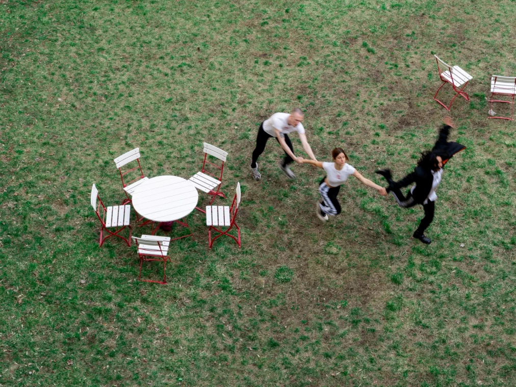 Drei Personen bewegen sich über eine Wiese und halten sich an den Händen. Die Szene ist aus der Vogelperspektive aufgenommen. Neben ihnen stehen mehrere weiße Gartenstühle und ein runder Tisch mit rotem Gestell.