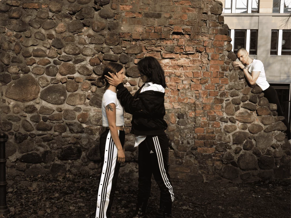 Drei Personen stehen im Freien vor einer alten Steinmauer. Zwei Personen stehen sich nahe gegenüber, während eine Person der anderen das Gesicht mit beiden Händen hält. Eine dritte Person lehnt seitlich auf der Mauer und beobachtet die Szene.