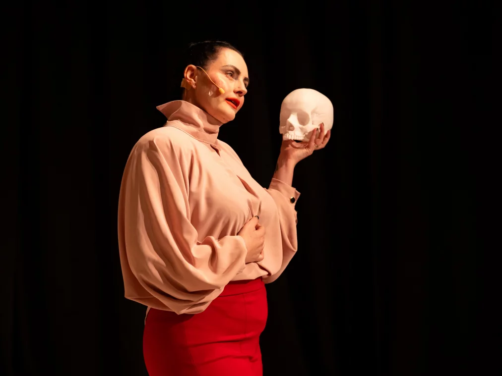 A person stands sideways on stage, holding a white skull at shoulder height while placing the other hand on their torso.