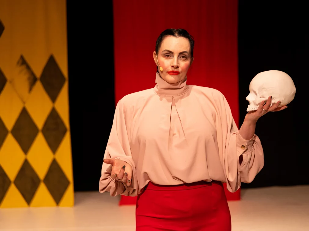 A person faces the audience, holding a white skull in their right hand. Their left hand is open and directed forward.