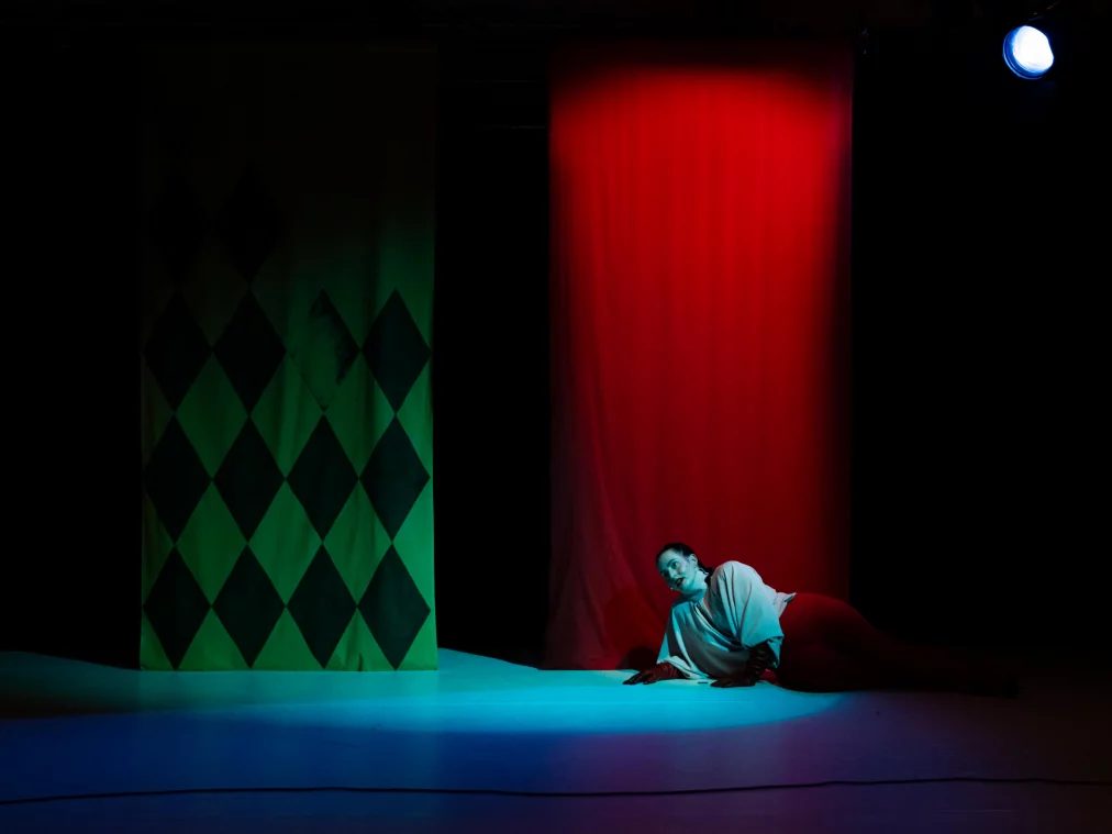 A person lies on their side on the stage floor in colored light, supporting themselves on one arm. Large red and green fabric panels hang behind them.