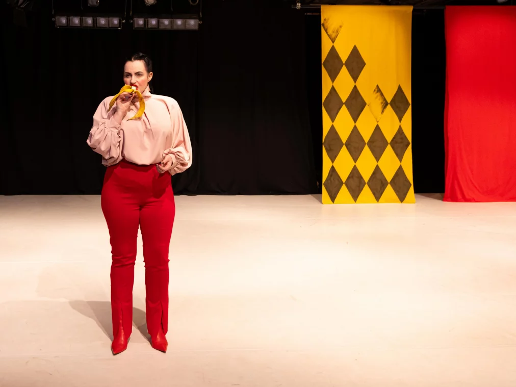 A person stands on stage eating a banana. Two large fabric panels with diamond patterns hang in the background.