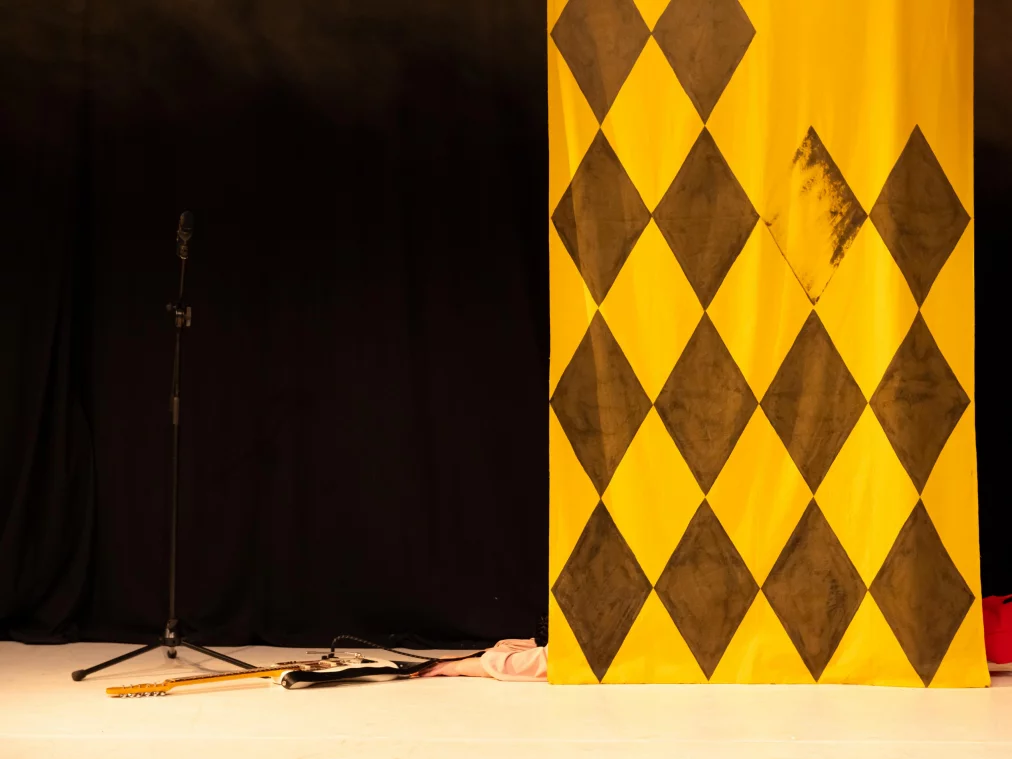 A person lies partly hidden behind a yellow fabric panel on the floor. An electric guitar and a microphone stand lie nearby.