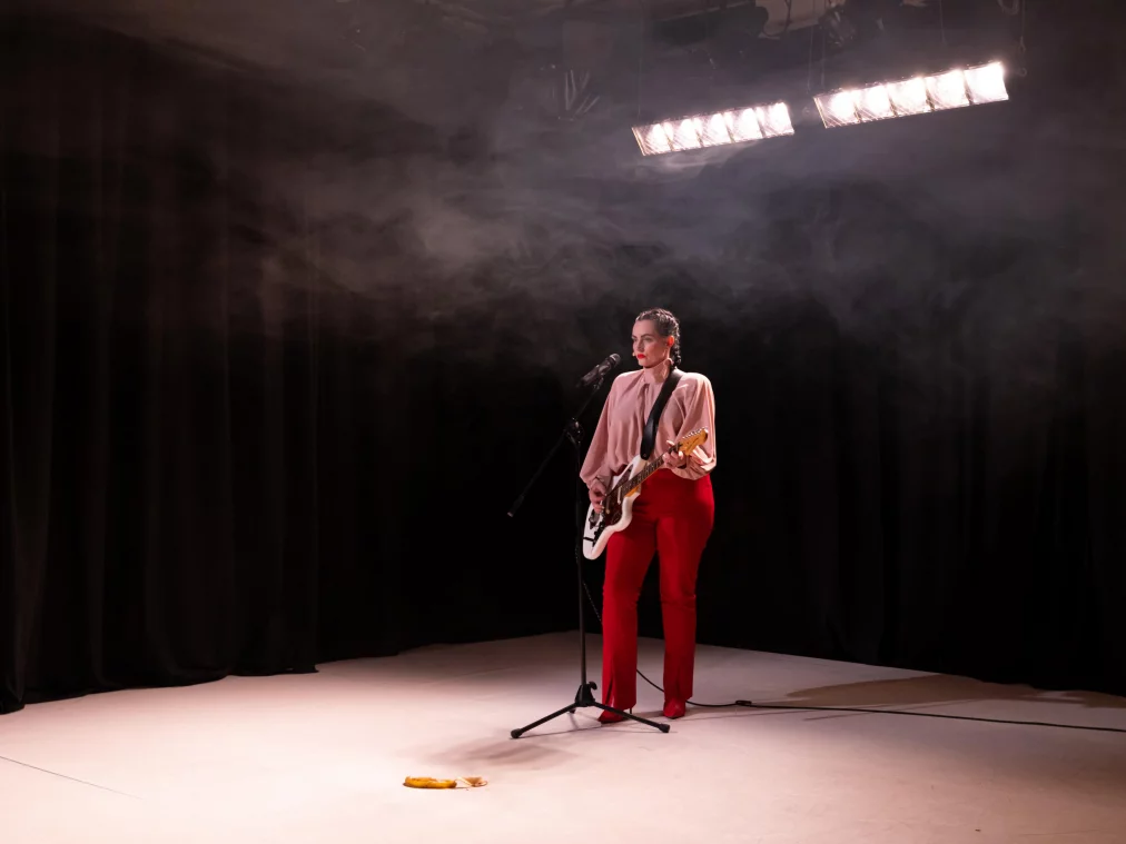 A person stands under bright stage lights with an electric guitar at a microphone, playing. A banana peel lies on the floor.