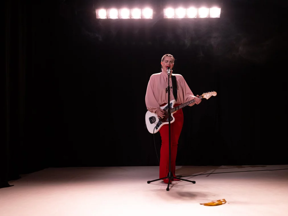 A person sings into a microphone while playing an electric guitar. Bright stage lights are visible above, and a banana peel lies on the floor in front of them.