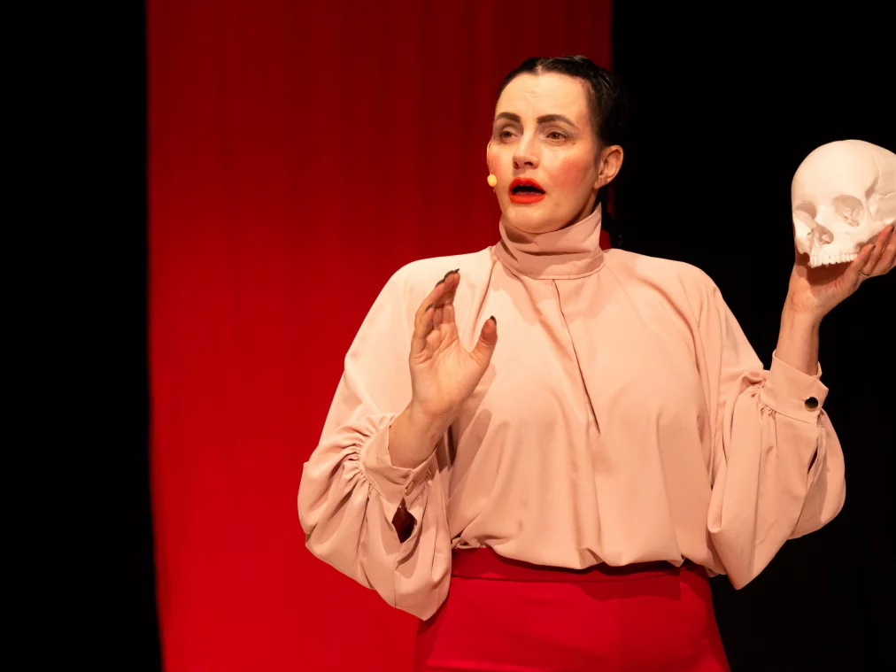 A person stands on stage in front of a red backdrop, holding a white skull in their right hand while their left hand is raised in a gesture.