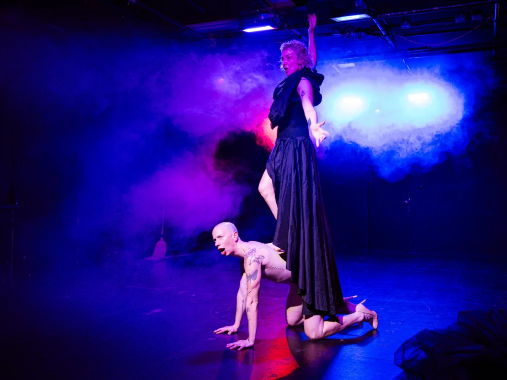 Zwei Personen bewegen sich über eine Bühne unter blau-violettem Bühnenlicht.