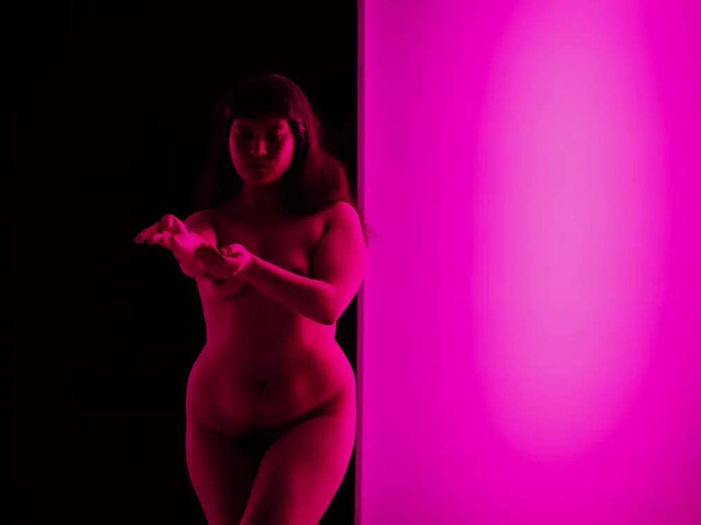 Eine Person steht neben einer leuchtend pinken Wandfläche im dunklen Bühnenraum. Der Körper ist von pinkem Licht beleuchtet, während der Hintergrund schwarz bleibt. Die Person hält eine Hand vor sich und betrachtet sie.