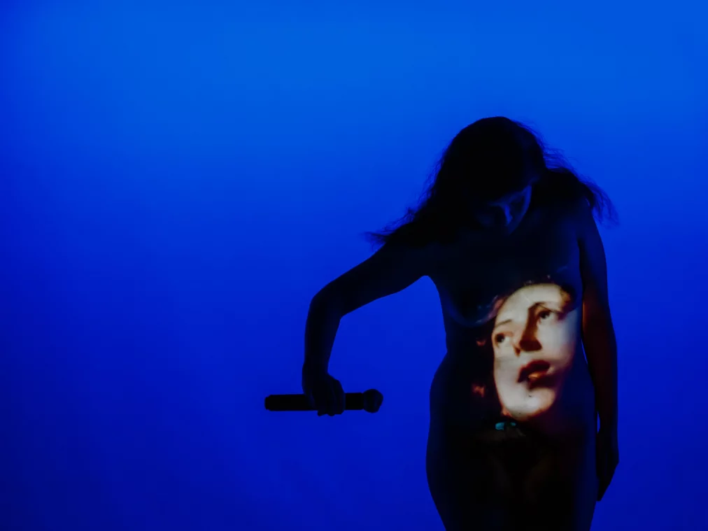 Eine Person steht in einem blau beleuchteten Raum und hält ein Mikrofon locker in der Hand. Auf den Oberkörper der Person ist das projizierte Bild eines Gesichts zu sehen, das sich über Brust und Bauch legt. Die Person blickt nach unten auf die Projektion.