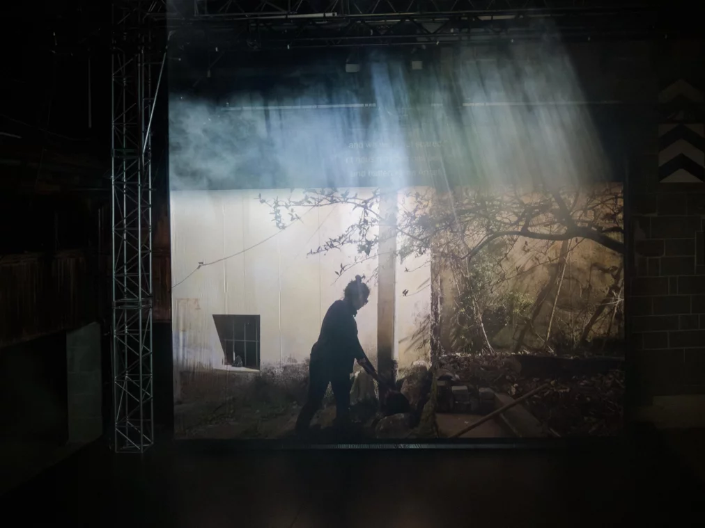Eine große Videoprojektion zeigt die Silhouette einer Person, die sich in einem Hof oder Garten bewegt. Lichtstrahlen fallen von oben durch Bühnennebel und erzeugen sichtbare Lichtbündel über der Projektion.