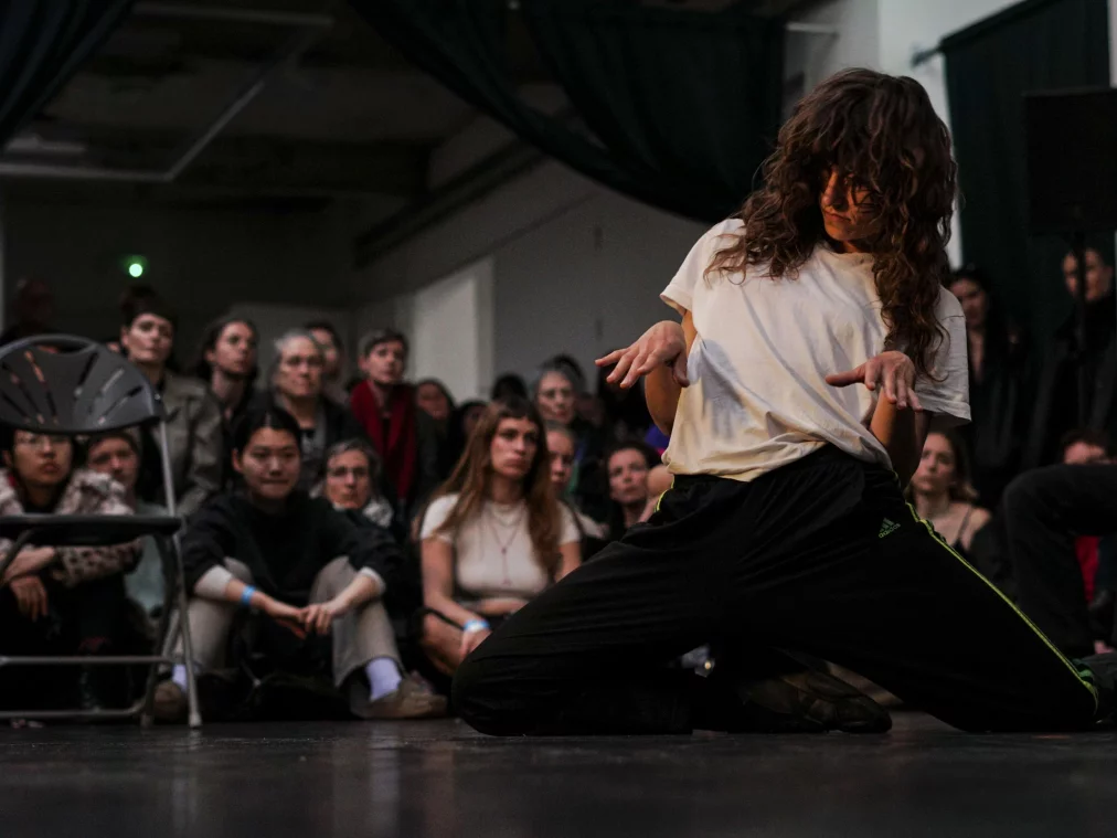Eine Person kniet breitbeinig auf dem Bühnenboden und hält die Hände mit angewinkelten Fingern vor dem Körper. Hinter ihr sitzt ein Publikum und verfolgt die Performance aufmerksam.