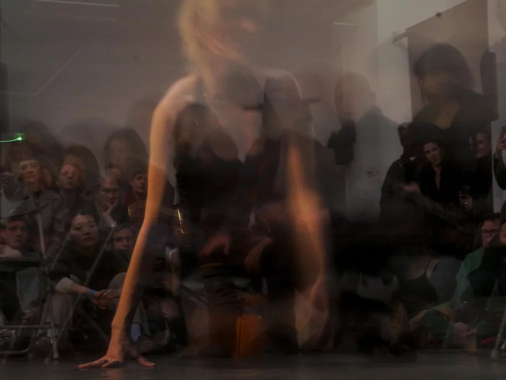 Eine Person bewegt sich unscharf und schnell über den Bühnenboden, sodass der Körper verschwimmt. Im Hintergrund sitzt ein Publikum dicht gedrängt auf Stühlen und beobachtet die Szene.