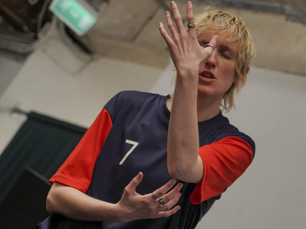 Eine Person hält eine Hand vor das Gesicht und blickt durch die gespreizten Finger. Die andere Hand ist vor dem Oberkörper positioniert, während der Hintergrund von einer industriellen Innenarchitektur geprägt ist.