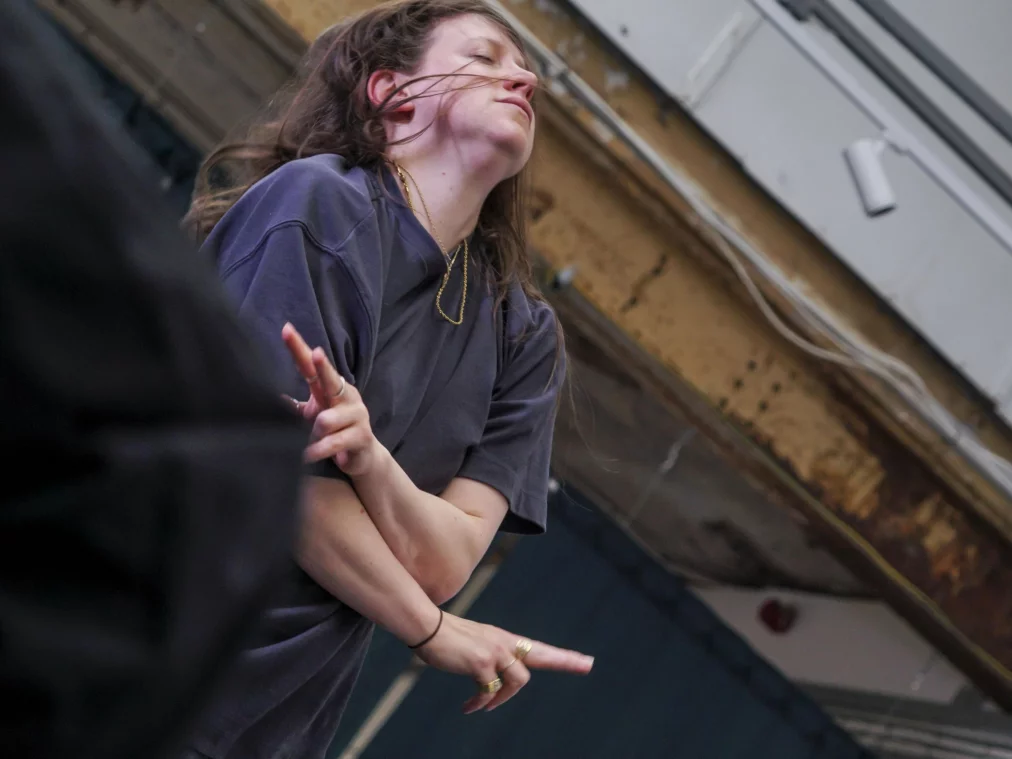 Eine Person mit geschlossenen Augen neigt den Kopf leicht nach hinten und bewegt die Hände in einer gestischen Bewegung. Der Raum wirkt roh, mit sichtbaren Kabeln und einer industriellen Decke.