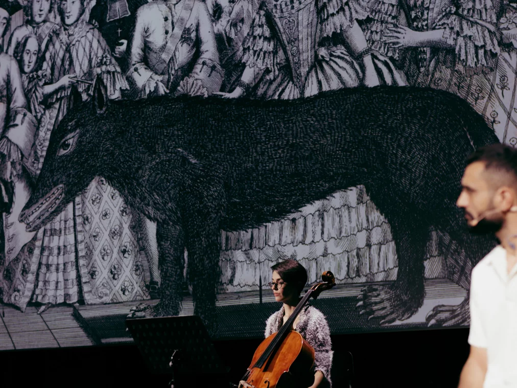 Eine Person sitzt mit einem Cello vor einem Notenständer auf der Bühne. Im Hintergrund ist eine großformatige Projektion eines schwarz-weißen Bildes zu sehen, das ein großes Tier zeigt, während eine weitere Person unscharf im Vordergrund steht.