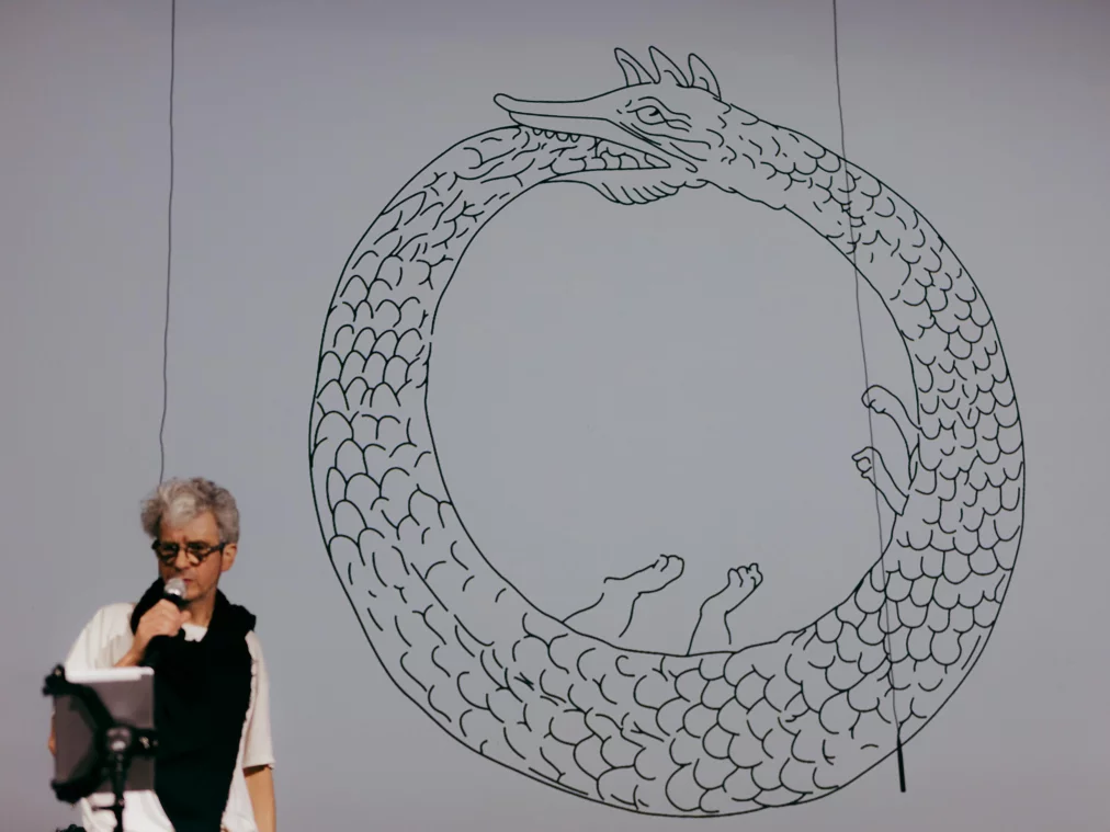 Eine Person steht links neben einem Mikrofon und liest von einem Tablet oder Blatt. Hinter ihr ist eine großflächige Projektion einer gezeichneten Schlange zu sehen, die sich kreisförmig selbst in den Schwanz beißt und in deren Innerem angedeutete Körperteile erscheinen.