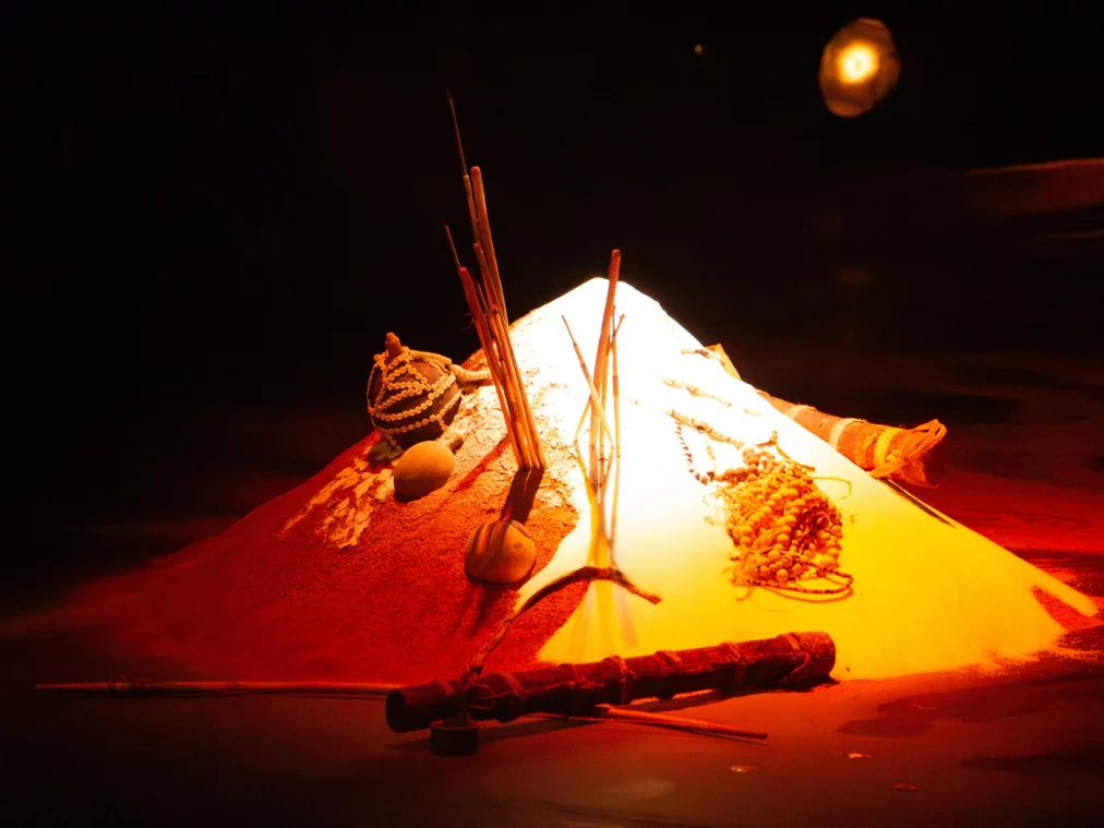 Ein beleuchteter Sandhaufen liegt in dunkler Umgebung und ist mit verschiedenen Objekten arrangiert: mehrere aufrecht stehende Räucherstäbchen, Perlenketten, kleine Gefäße, Stäbe und weitere rituell wirkende Gegenstände. Warmes, rötlich-gelbes Licht hebt die Mitte des Haufens hervor und lässt die Umgebung im Dunkeln verschwinden.