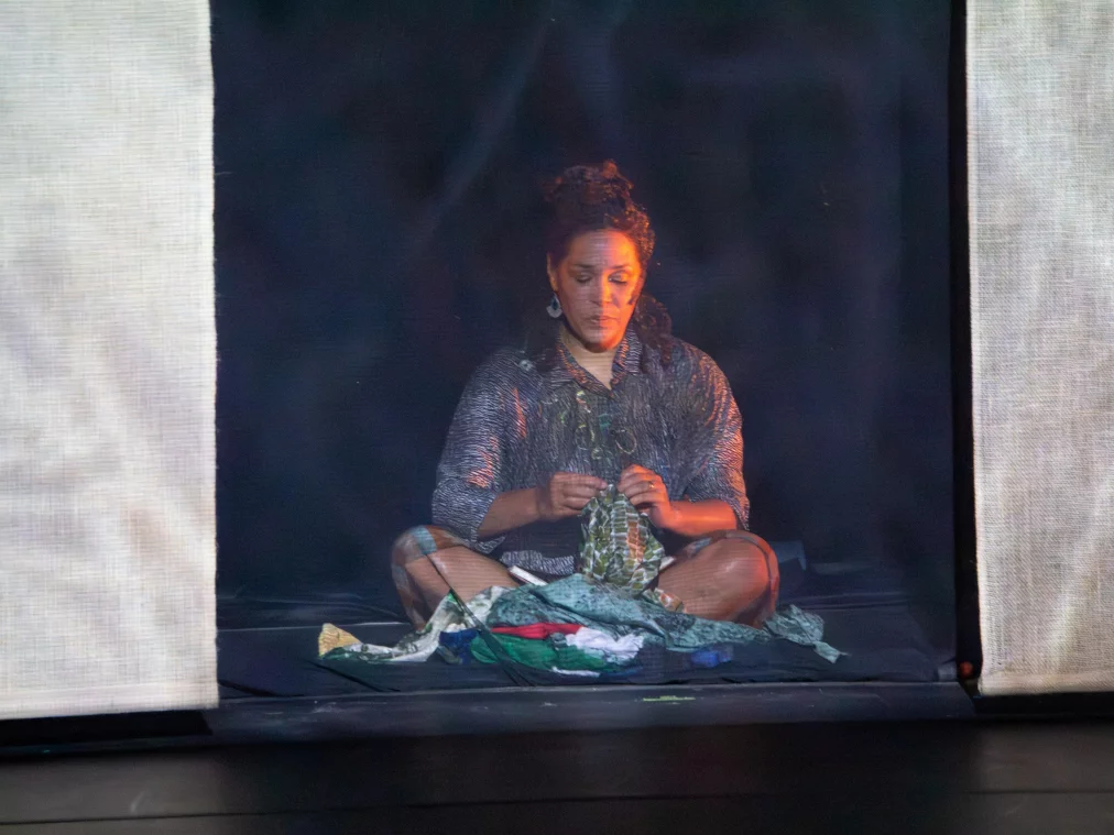 Eine Person sitzt auf dem Boden in einem von Stoffbahnen gerahmten Bühnenraum und arbeitet mit einem Stück Stoff oder Kleidungsstück in den Händen. Die Haltung ist ruhig und konzentriert, das Licht fällt weich von vorne auf die Person, während der Hintergrund dunkel bleibt.