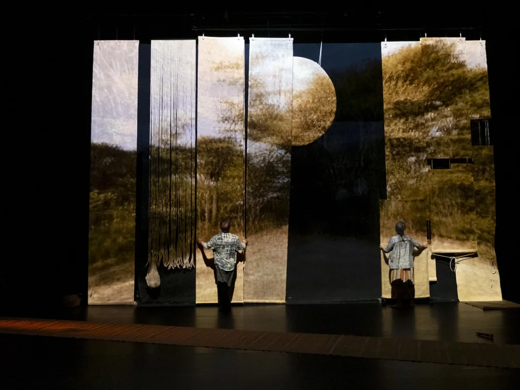 Zwei Personen stehen auf einer Bühne vor hohen, schmalen Stoffbahnen, auf die eine Landschaft mit Bäumen projiziert ist. Die Stoffbahnen hängen nebeneinander und bilden eine fragmentierte Bildfläche, während die Personen jeweils eine Bahn berühren oder halten.