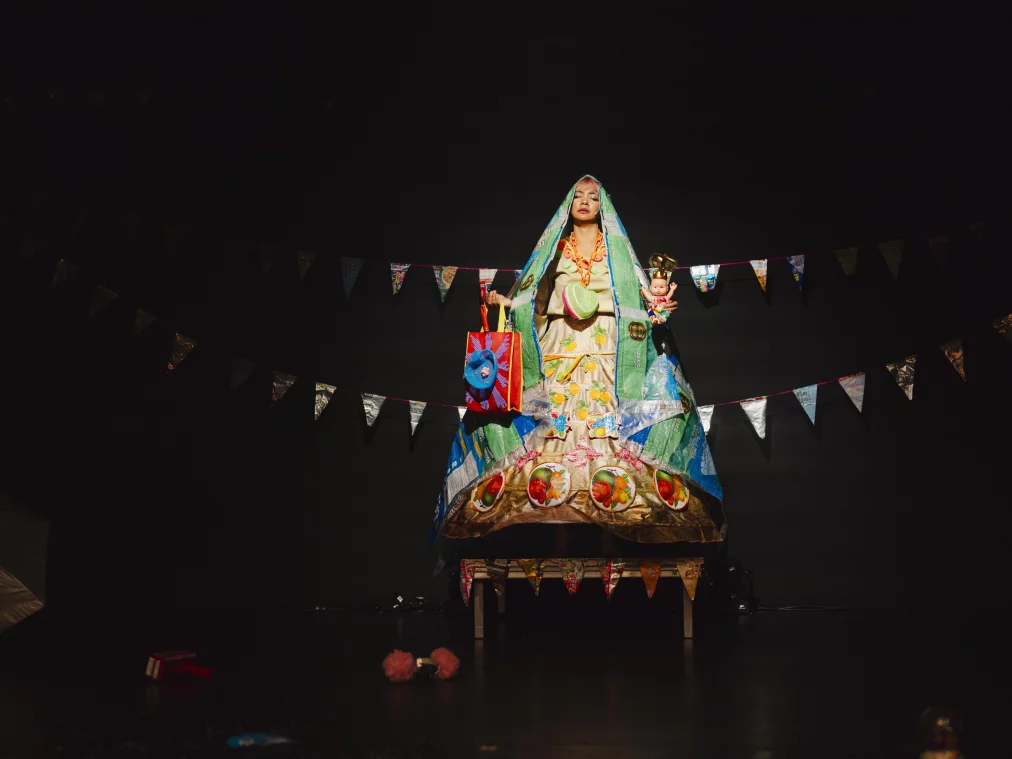 Eine Person steht erhöht auf einer Bühne in einem voluminösen, aus recycelten Materialien gefertigten Kleid, das sich glockenförmig ausbreitet. Die Person hält eine Tasche und eine Puppe, während hinter ihr Wimpelketten gespannt sind. Einzelne Gegenstände liegen auf dem Bühnenboden im Vordergrund, das Licht konzentriert sich auf die Person und lässt den Raum ansonsten dunkel.