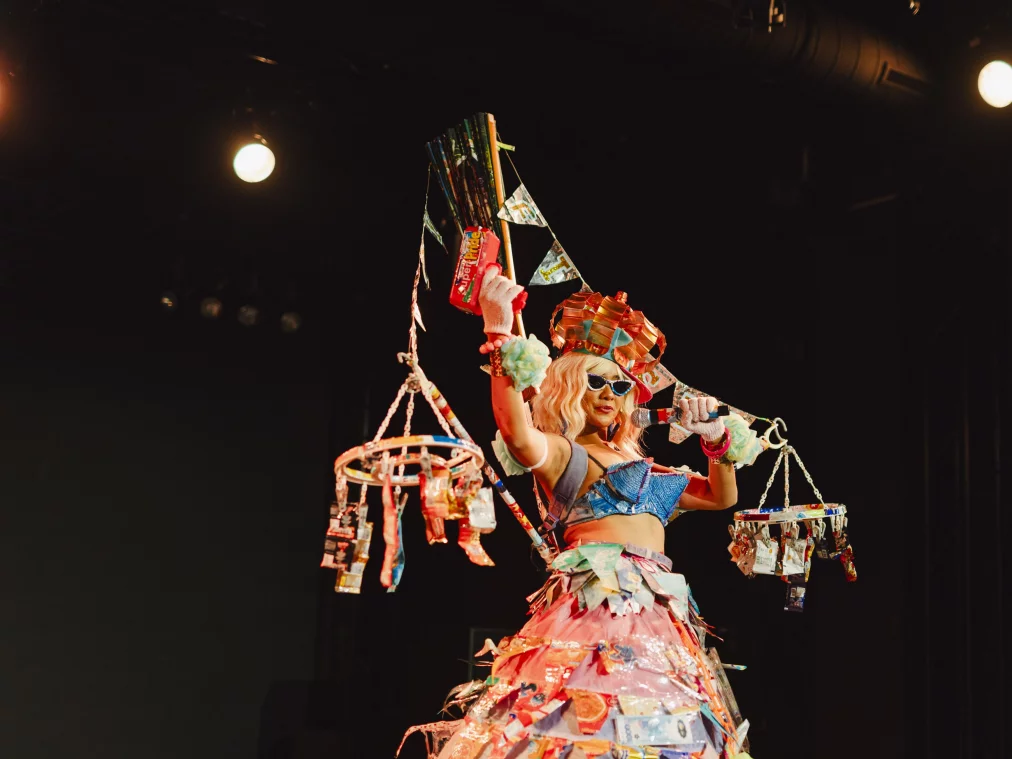 Eine Person in einem farbenfrohen, aus vielen Materialien zusammengesetzten Bühnenkostüm hält ein Mikrofon und hebt einen Arm. Das Kostüm besteht aus mehreren Schichten und ist mit kleinen Objekten und Verpackungen verziert, die auch an zwei seitlich hängenden, mobilen Konstruktionen befestigt sind. Die Szene ist von warmem Bühnenlicht beleuchtet, der Hintergrund bleibt dunkel.