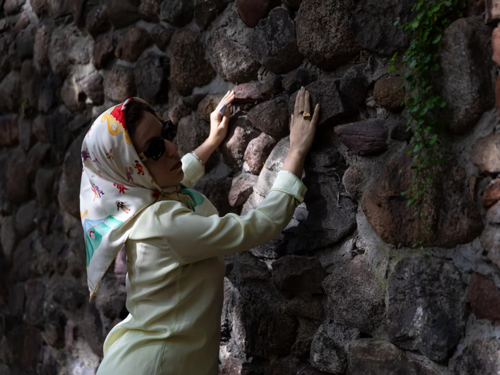 Eine Person steht seitlich vor einer rauen Steinmauer und legt beide Hände flach auf die unebene Oberfläche. Sie trägt ein helles Outfit, ein gemustertes Kopftuch und eine Sonnenbrille. Zwischen den Steinen wachsen kleine grüne Pflanzen, während Licht und Schatten die Szene strukturieren.
