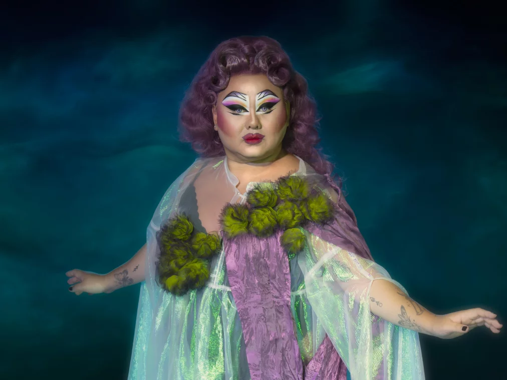 Eine Person mit auffälligem Drag-Make-up und violetter Lockenperücke steht vor einem dunklen, nebligen Hintergrund. Sie trägt ein schimmerndes, halbtransparentes Gewand in Grün- und Lilatönen mit floralen Applikationen und hält die Arme leicht ausgestreckt.