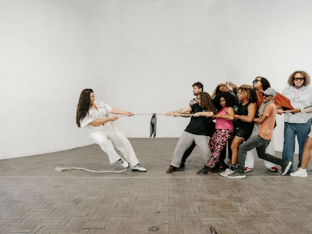 A group of people stands in a bright, empty room, pulling together on a rope in one direction. On the opposite side, a single person dressed in white holds the other end and leans back with their body weight. The body postures convey tension and resistance.