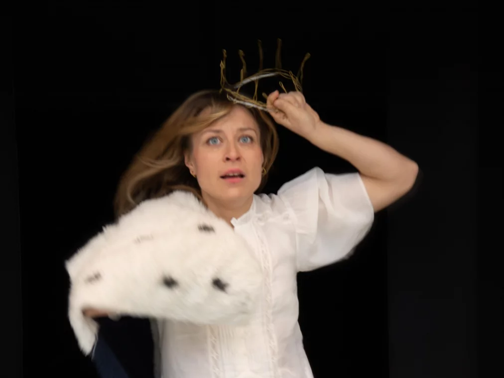 Eine Person mit kurzem hellem Haar bewegt sich vor einem schwarzen Hintergrund nach vorne. Sie trägt ein kurzes weißes Kleid, weiße kniehohe Stiefel und einen dünnen, kronenartigen Kopfschmuck aus Zweigen oder Draht. Über einem Arm liegt ein weißer Mantel mit dunklem Innenfutter. Die Person blickt mit geöffnetem Mund direkt nach vorne, während Haare und Kleidung in Bewegung geraten.