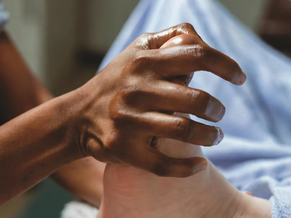 Eine Hand berührt sanft einen Fuß. Die Finger umschließen die Ferse, während der Fuß auf einem weichen, blauen Stoff liegt. Die Nahaufnahme betont Hautstruktur, Berührung und den Kontrast der Hauttöne.