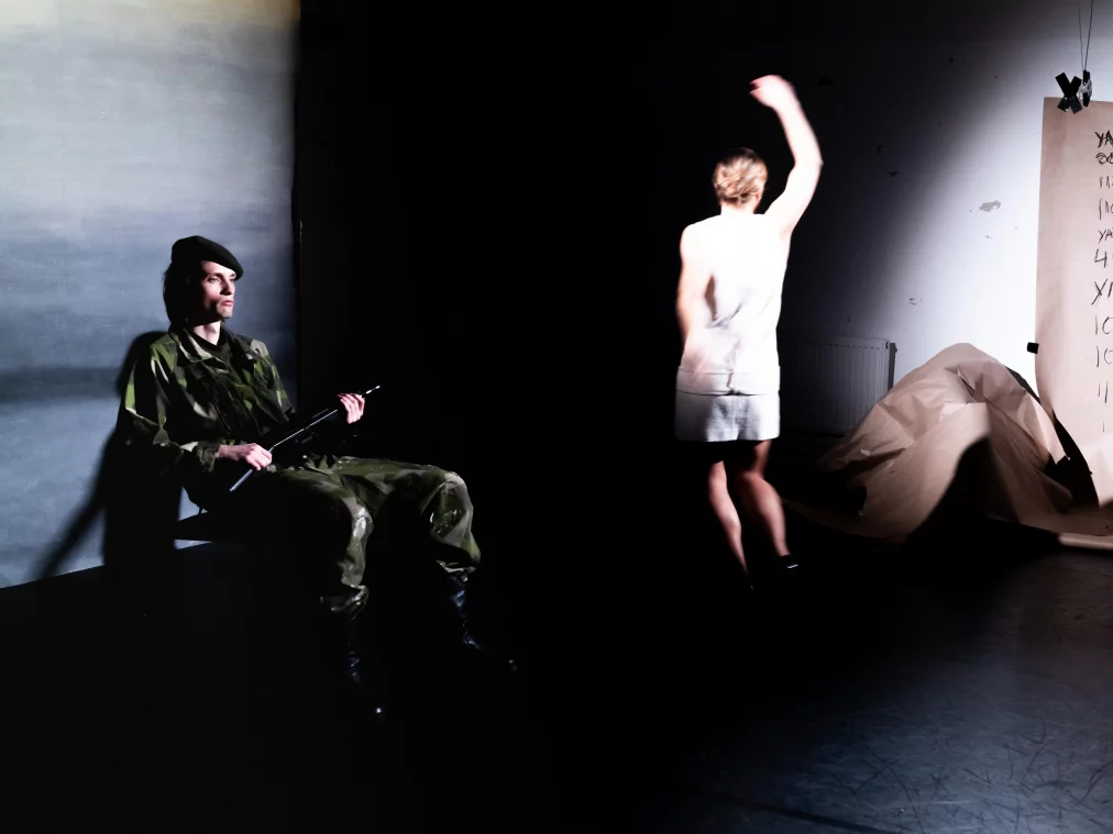 Theatre scene in a dark space. On the left, a person in camouflage clothing and a dark beret sits on a chair holding a weapon. In the centre, a person in a white dress dances with one arm raised, back to the camera, illuminated by a spotlight. On the right, a large paper poster with handwritten numbers and the word "YANKEE" hangs from above; crumpled paper lies on the floor in front of it.