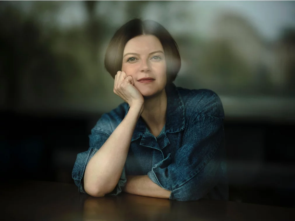 Helen Hester sitzt an einem Tisch und stützt das Kinn auf die verschränkten Hände. Sie trägt eine Jeansjacke und blickt nachdenklich in die Kamera. Im Hintergrund sind durch ein Fenster verschwommen Bäume zu sehen.