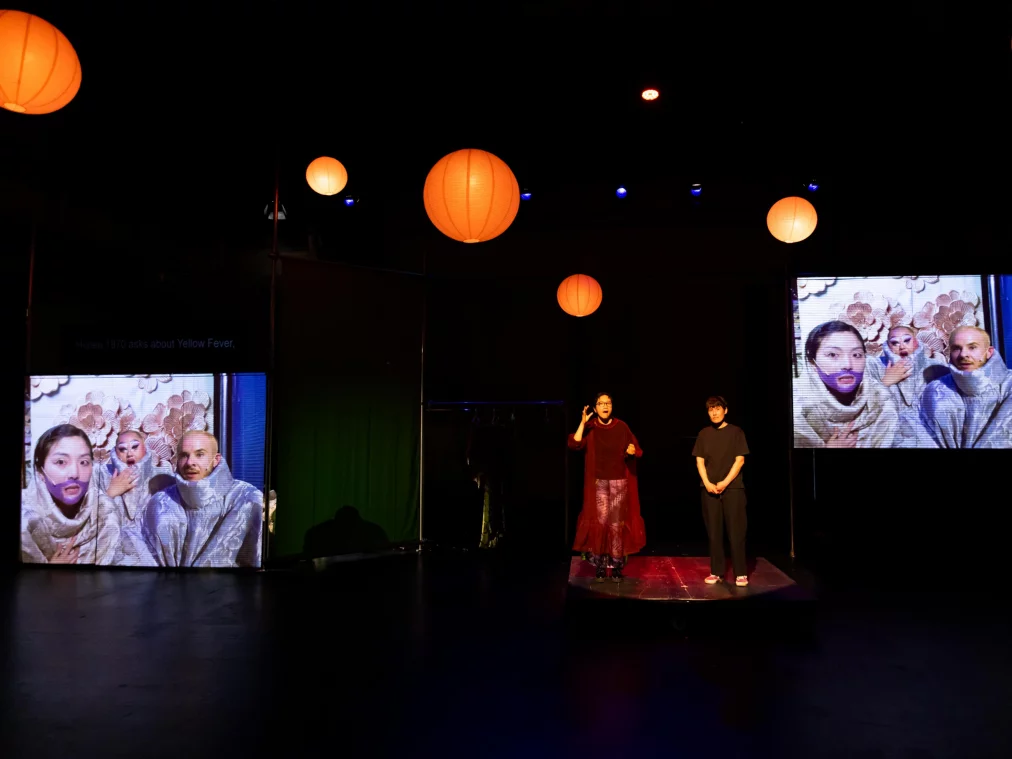 Wide theatre stage with orange paper lanterns. Two people stand centrally on a raised platform; one is gesturing. They are flanked by two large video projections, each showing three people in elaborate floral costumes and headdresses. The left projection displays the text "Hana: 1970 asks about Yellow Fever".
