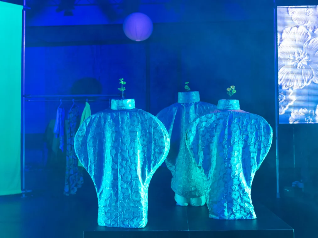 Three vase-shaped stage sculptures with a textured surface stand on a plinth, bathed in blue light. Each sculpture is topped with a small green plant. In the background, a LED screen showing a floral pattern is visible on the right, a costume rack on the left.
