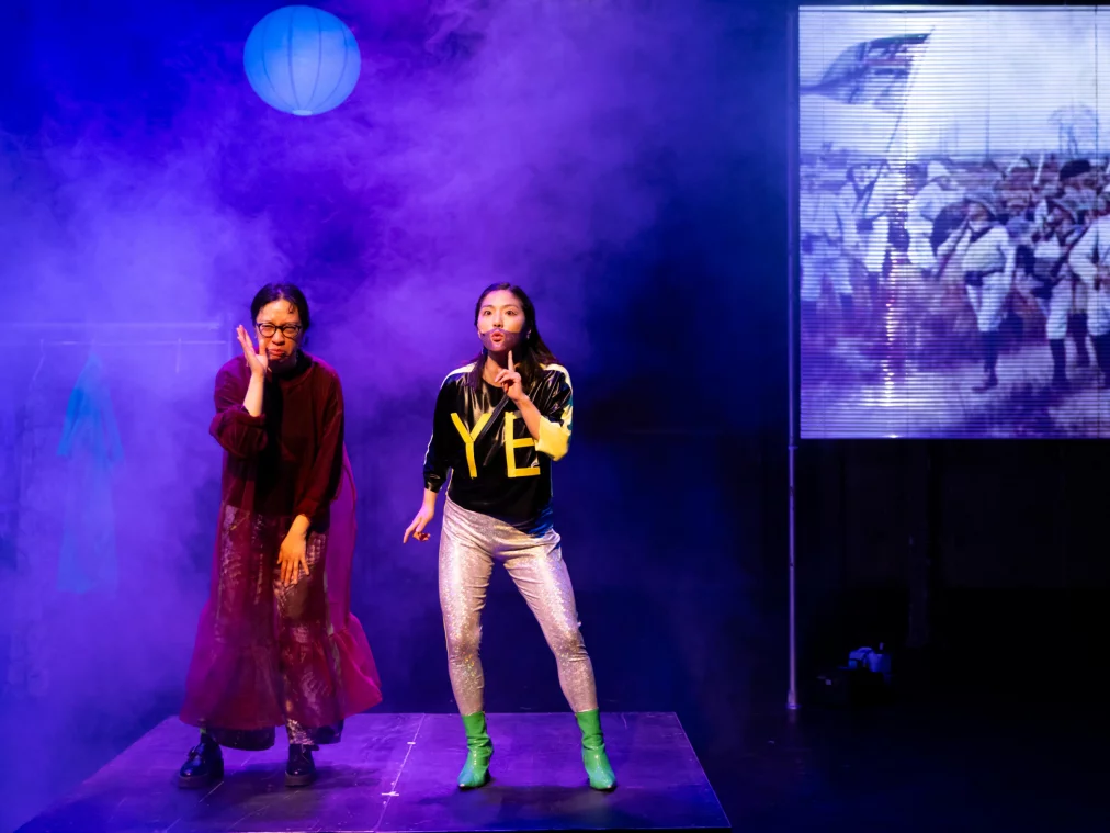 Two people on a stage filled with blue theatrical haze. The person on the left wears a dark red coat and glasses. The person on the right wears a black top with yellow lettering "YE", silver trousers and green boots, and holds up one finger. In the background, a blue paper lantern and an LED screen showing a historical painting of marching soldiers.