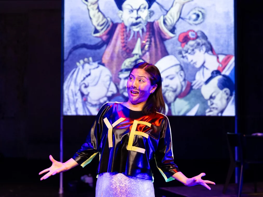 A person stands in the foreground of a dark stage with arms spread wide. They wear a black top with the yellow letters "YE", a glittery mini skirt, and have purple glitter on their face. In the background, a projection displays a historical caricature illustration.