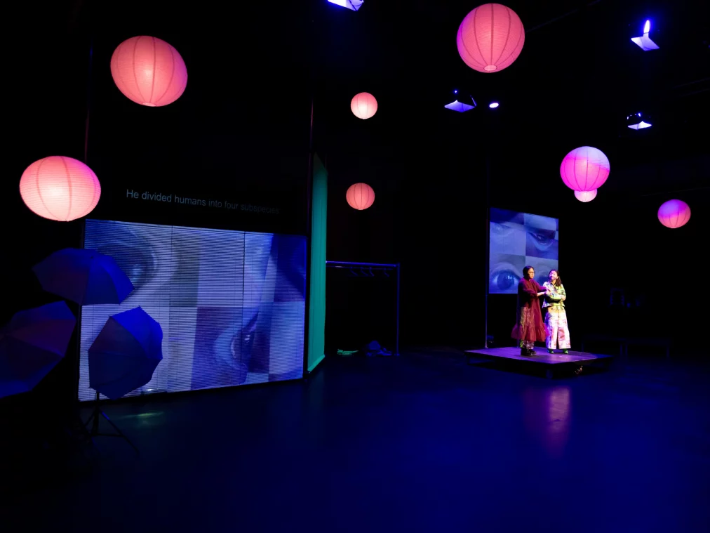 Wide theatre stage bathed in blue light with numerous pink paper lanterns. Two people stand on a raised platform right of centre. Two LED screens are visible behind them: one showing umbrellas, the other an eye motif. The left screen displays the text "He divided humans into four subspecies." Blue umbrellas are visible in the left foreground.