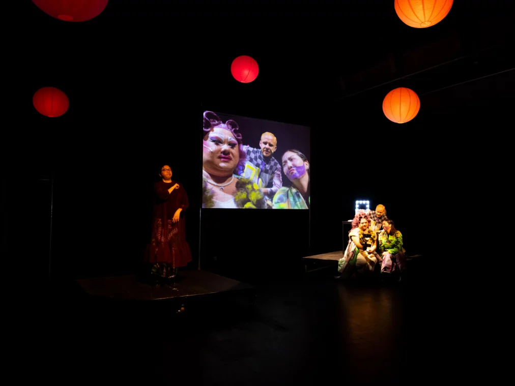 Dark theatre stage with red and orange paper lanterns. On the left, a person in a dark red dress. In the centre, a projection screen showing three people in expressive make-up and colourful costumes. On the right, three people sit on a raised platform next to an illuminated mirror.