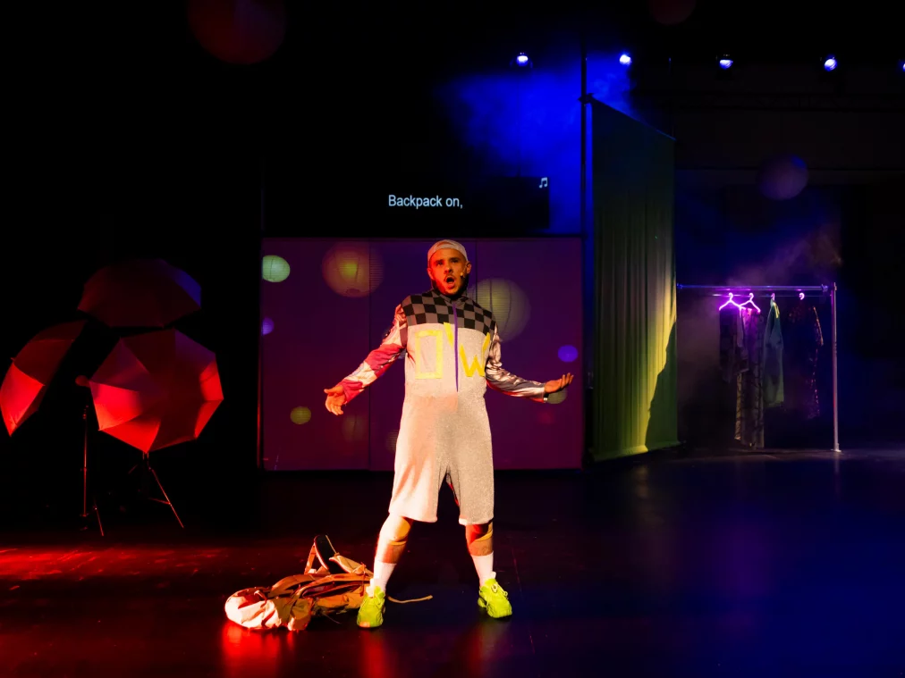 A person stands centre stage on a dark stage, singing. They wear white shorts, a checked top with yellow lettering, white knee socks and neon green sneakers. A backpack lies at their feet. In the background, a video projection reads "Backpack on,", a costume rack and red umbrellas are visible.
