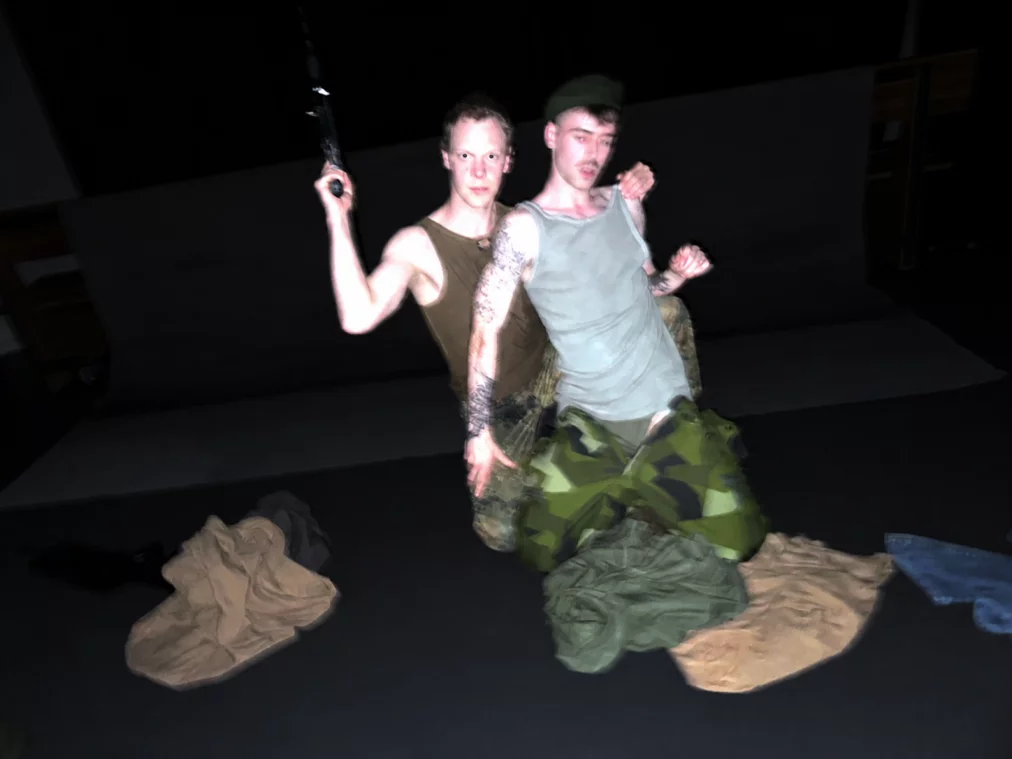Two people in tank tops kneel on a dark stage, surrounded by military clothing scattered on the floor. One person holds a weapon raised, both looking into the camera. The image is slightly blurred.