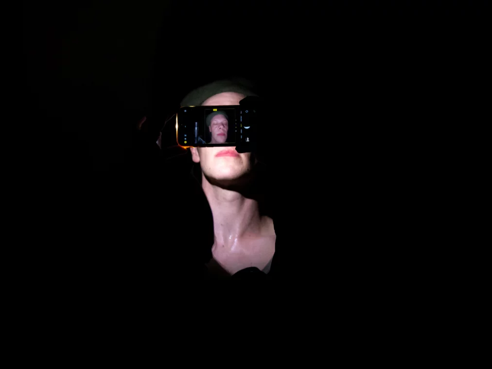 Eine Person hält ein Smartphone vor ihr Gesicht, sodass das Display die Augen verdeckt. Auf dem Display ist das Gesicht der Person zu sehen. Dunkler Hintergrund.