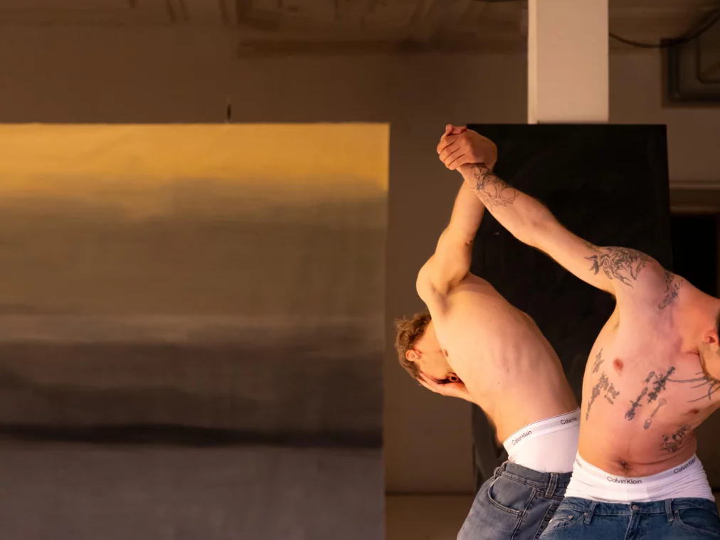 Zwei Personen mit Oberkörper-Tätowierungen in Jeans und weißer Unterwäsche führen auf einer Theaterbühne eine akrobatische Bewegung aus – die Oberkörper sind stark nach hinten gebeugt, die Hände greifen ineinander. Im Hintergrund eine großformatige Landschaftsmalerei.